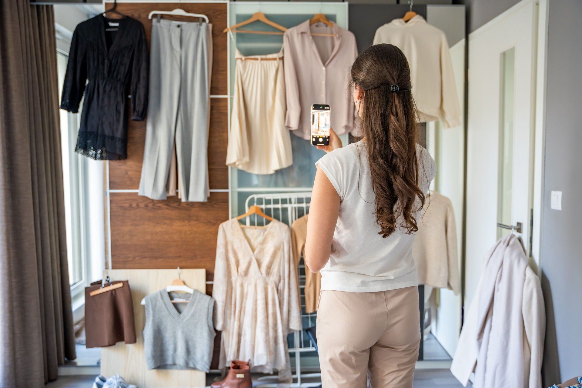 Young woman photographs clothing hanging on a wardrobe to create content for social media or a personal fashion catalog. Digital platforms and online presence shape modern fashion trends