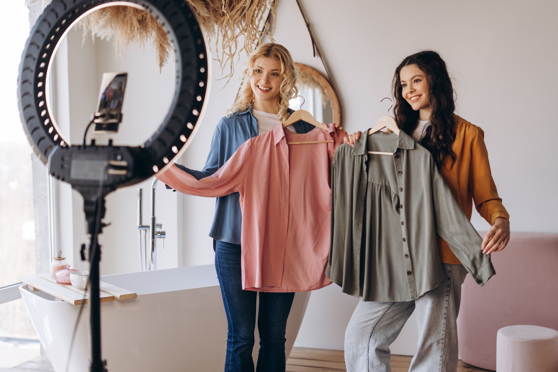 Happy two girlfriends recording video vlog while demonstrating shirts. using phone on tripod